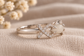 A silver ring featuring a central infinity symbol paved with small clear stones and a milky white oval stone, resting on a textured beige fabric background with dried flowers.